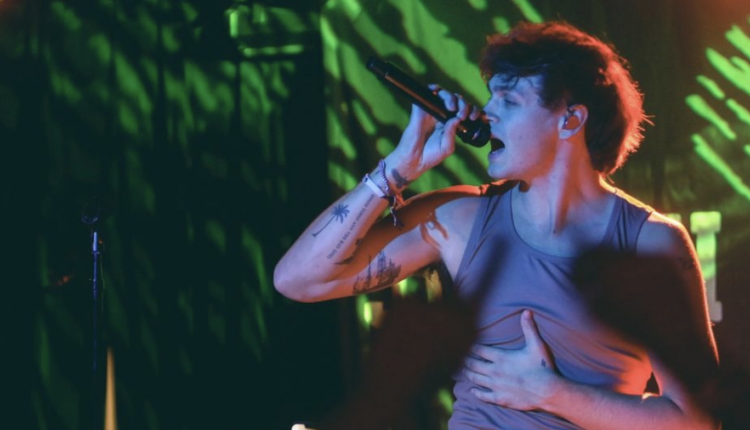 Jonah posing to the left while holding onto the microphone. Their are bright green lights behind him.